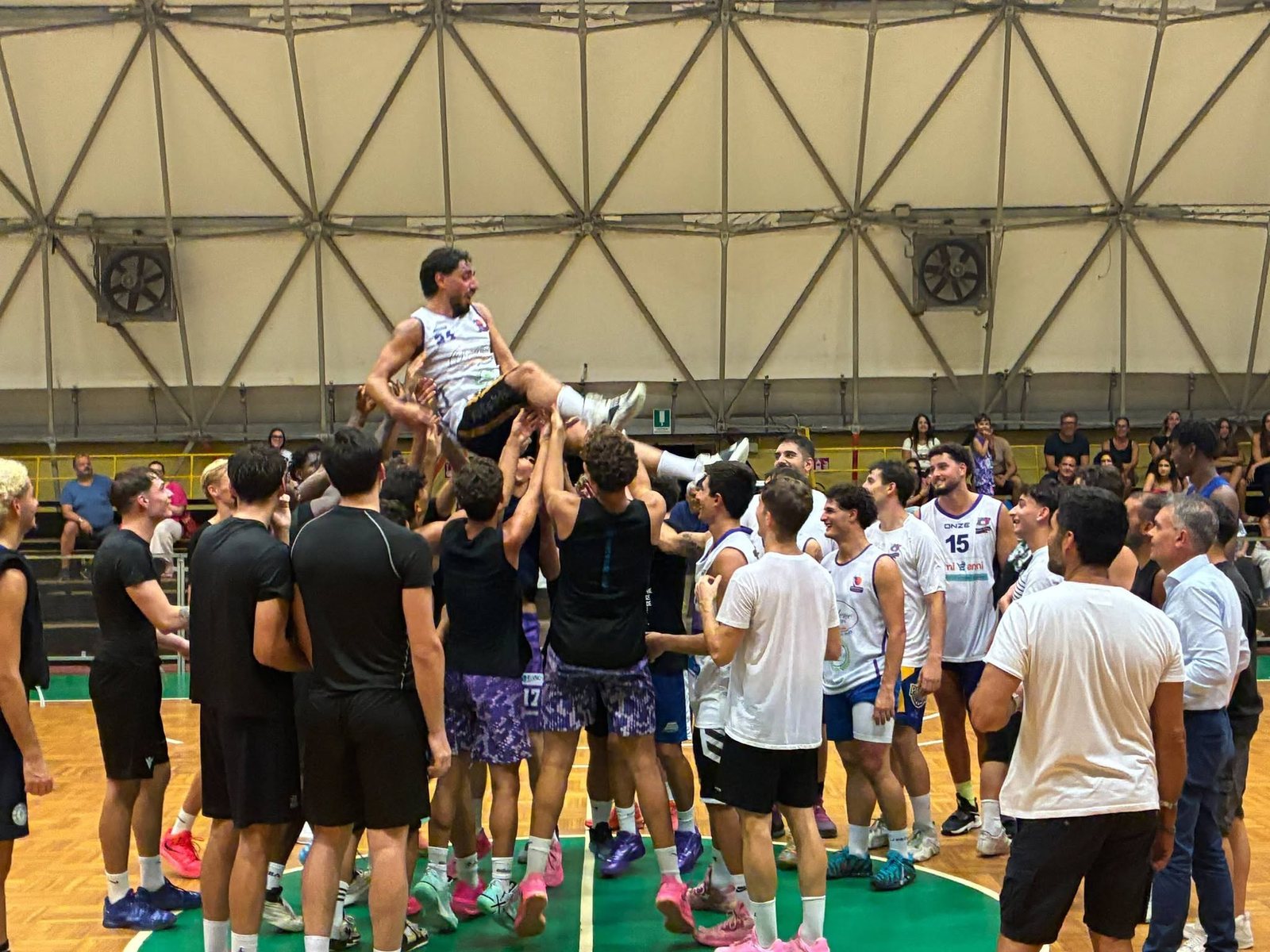 Un giocatore in maglia bianca viene sollevato in aria da un gruppo di compagni di squadra a centrocampo del Palamangione di Gioia Tauro, circondato da altri giocatori in maglia bianca e nera; sullo sfondo il pubblico sugli spalti.