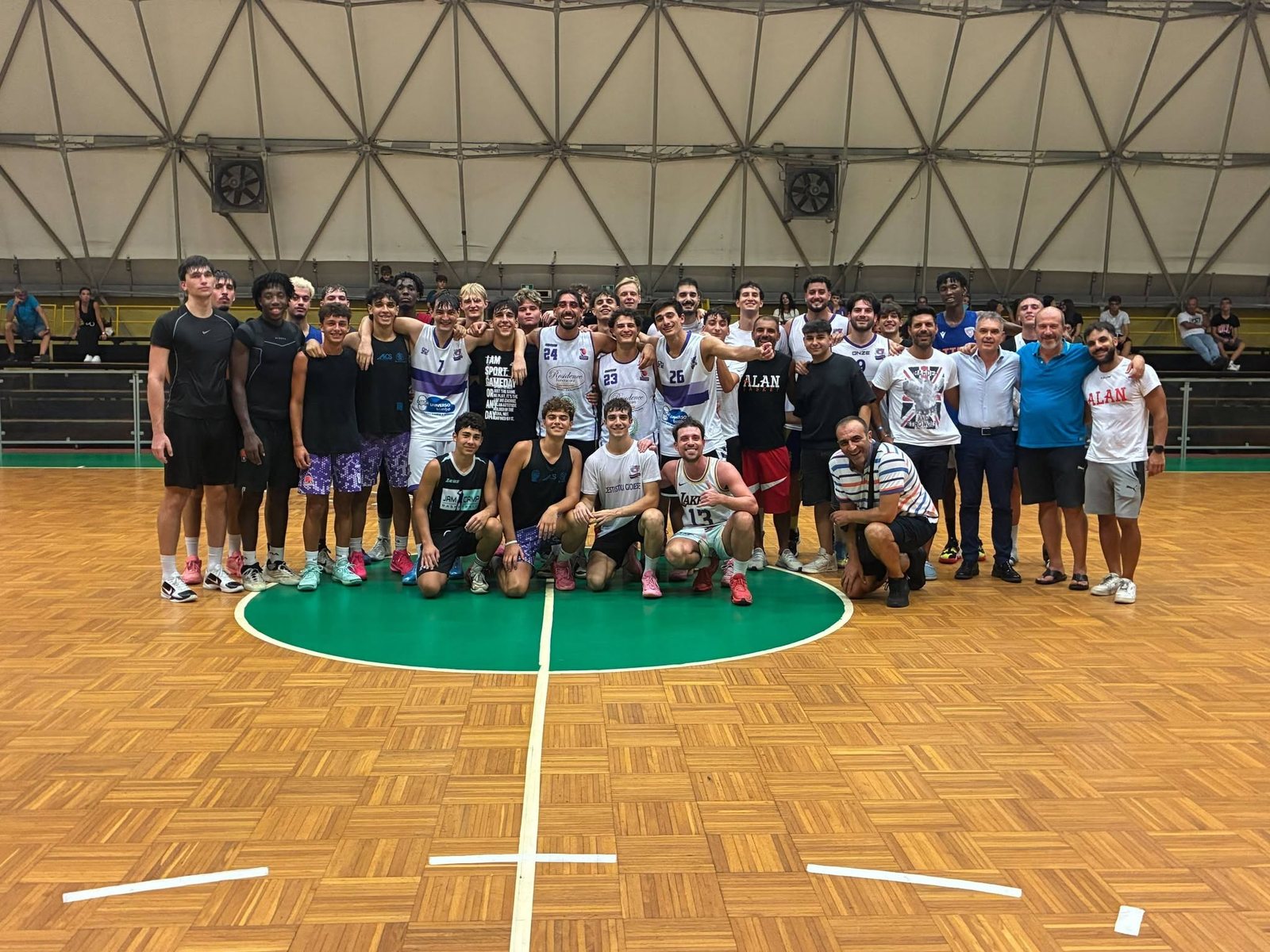 Un ampio gruppo di giocatori, alcuni in maglia bianca e altri in maglia scura, posa insieme al centro del campo del Palamangione di Gioia Tauro, in piedi e in ginocchio sul cerchio di centrocampo verde, con il pubblico visibile sugli spalti alle spalle.